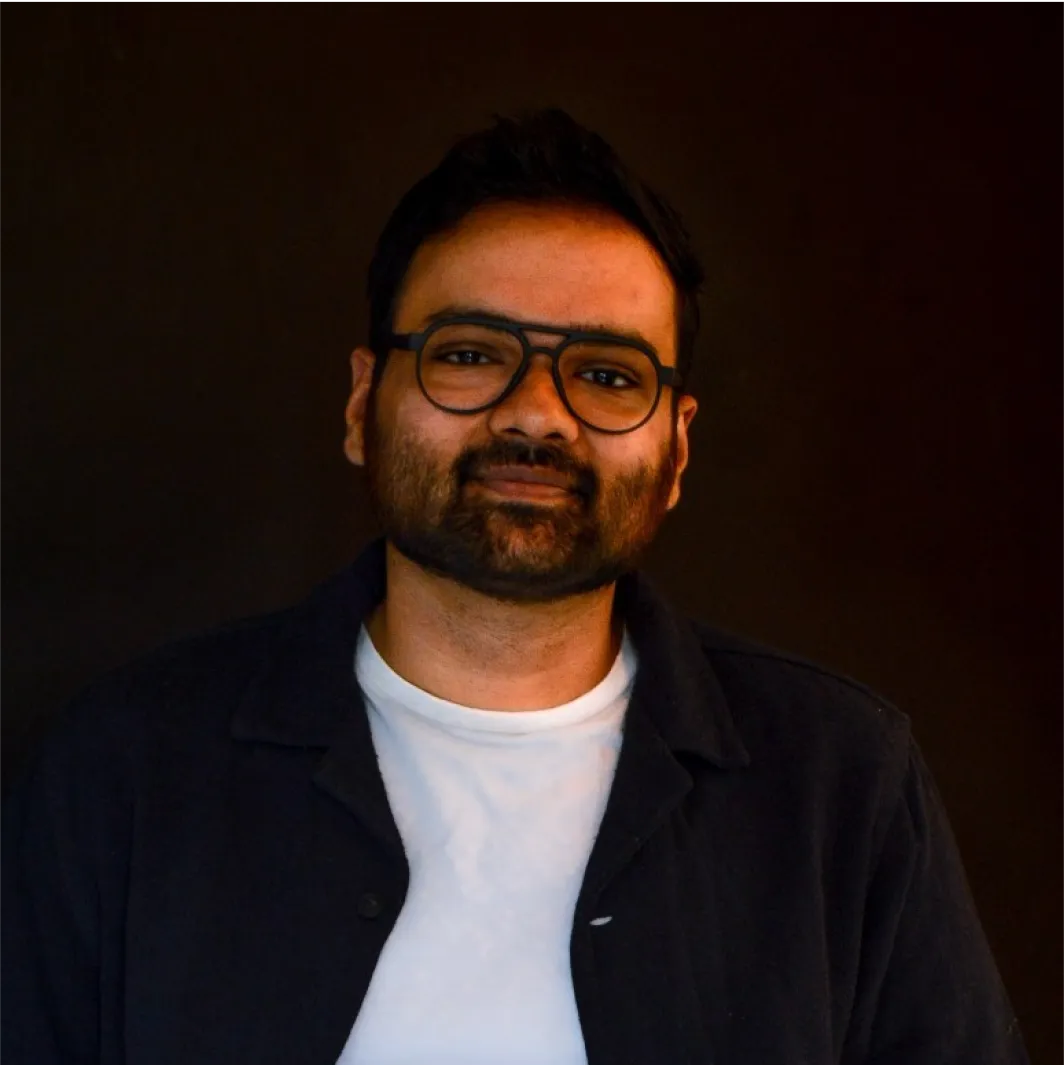 Man with dark hair, beard, and glasses wearing a white shirt and dark jacket against a dark background.